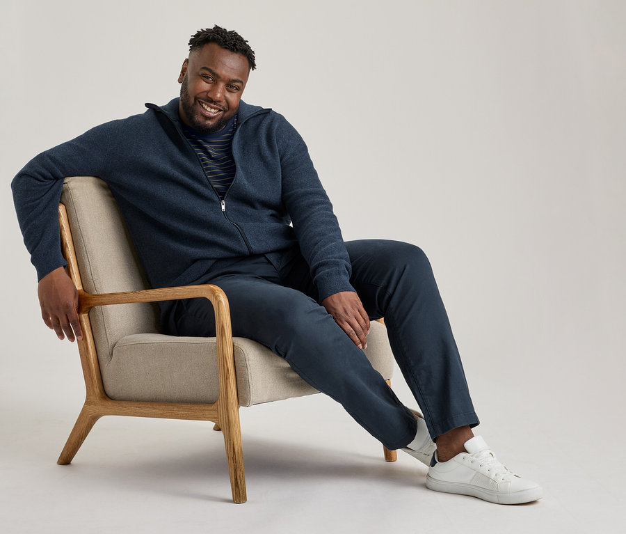 Lächelnder Mann sitzt in einem Sessel, trägt eine marineblaue Reißverschlussjacke, ein gestreiftes T-Shirt, eine navy Chino-Hose und weiße Sneaker.