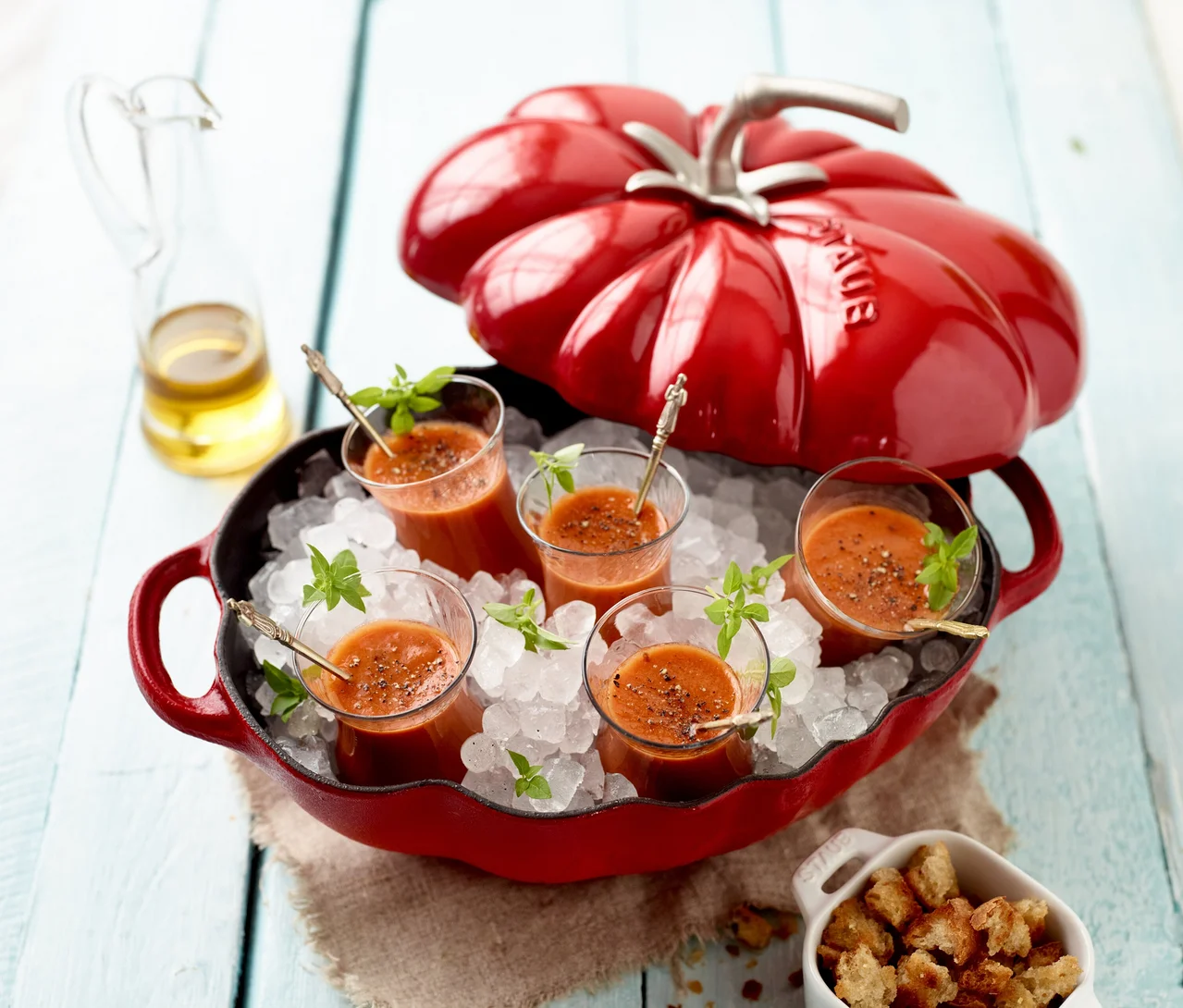 Roter STAUB Gusseisen-Bräter "Tomate" (ca. 25 cm) gefüllt mit Eis und fünf Gläsern Tomatensuppe, garniert mit Basilikum, daneben Olivenöl und Croutons.