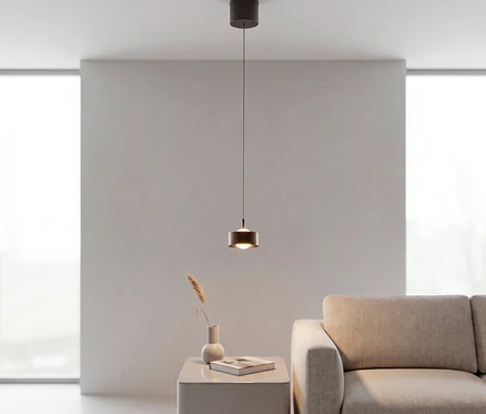 Eine moderne, dunkelbraune zylindrische Pendelleuchte hängt über einem hellgrauen Beistelltisch mit Vase und Buch, neben einem beigen Sofa in einem minimalistischen Raum mit großen Fenstern.