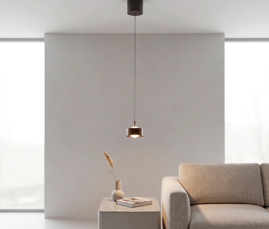 Eine moderne, dunkelbraune zylindrische Pendelleuchte hängt über einem hellgrauen Beistelltisch mit Vase und Buch, neben einem beigen Sofa in einem minimalistischen Raum mit großen Fenstern.