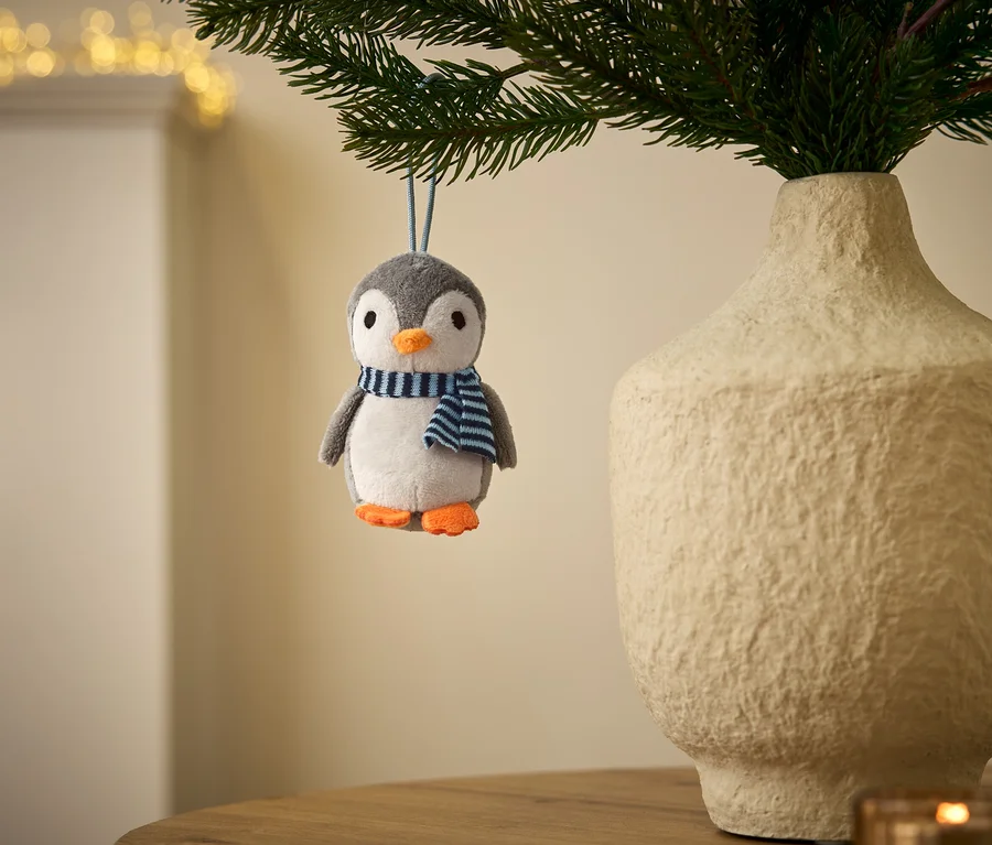 Ein grau-weißer Plüschpinguin-Anhänger mit orangefarbenem Schnabel und Füßen, der einen blau-weiß gestreiften Schal trägt, hängt an einem grünen Zweig. Daneben steht eine strukturierte, hellfarbene Vase mit Zweigen, im Hintergrund sind verschwommene Lichter zu sehen.
