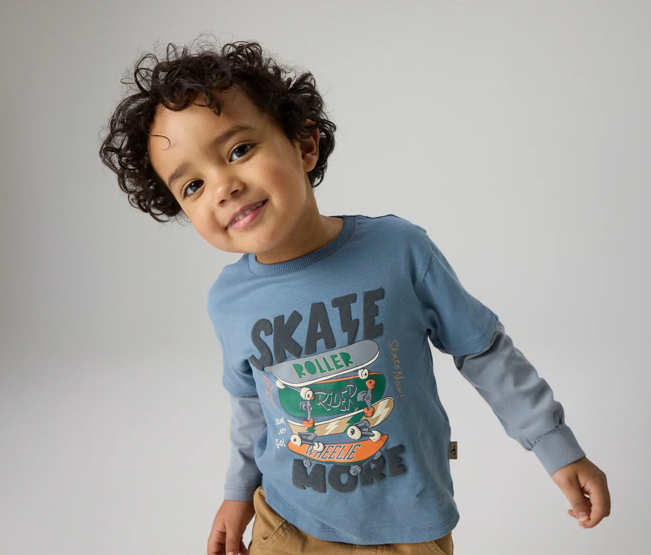 Lächelnder kleiner Junge mit lockigem dunklem Haar, der ein blaues Langarmshirt mit "SKATE"-Grafik und gestapelten Skateboards sowie braune Hosen trägt und in die Kamera blickt.