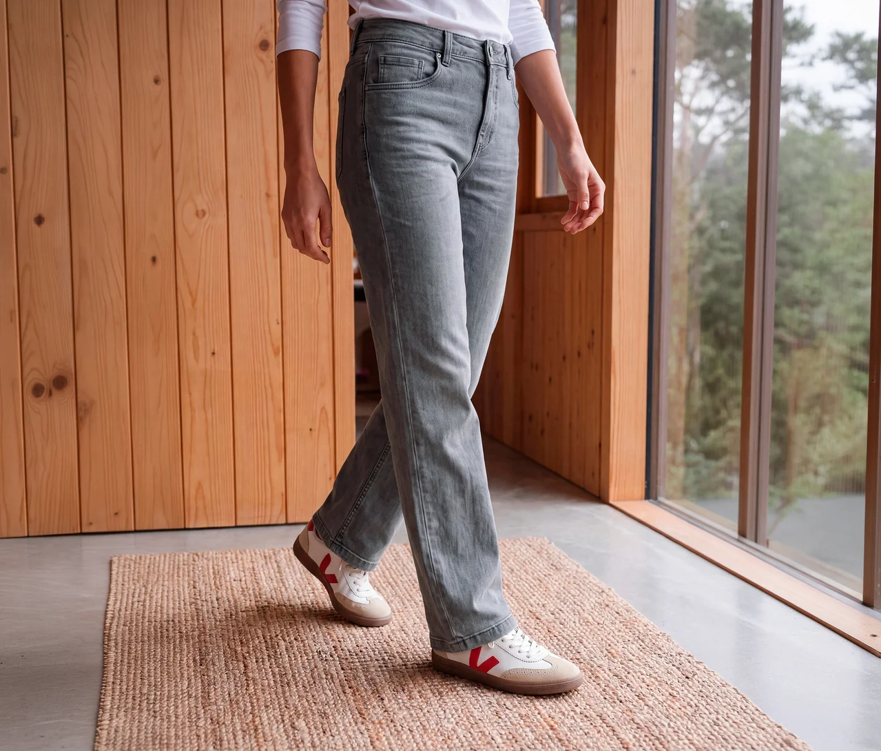 Eine Person geht, trägt eine helle graue CECIL Jeans, ein weißes Langarmshirt und weiße Sneaker mit roten Akzenten, auf einer gewebten Matte vor einer Holzwand und einem Fenster.