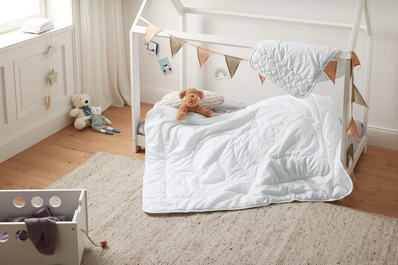Ein Kinderbett mit weißem Rahmen, dekoriert mit Wimpeln, mit einer weißen irisette® greenline Kinder-Steppdecke und einem Kissen. Auf dem Bett liegt ein Teddybär.