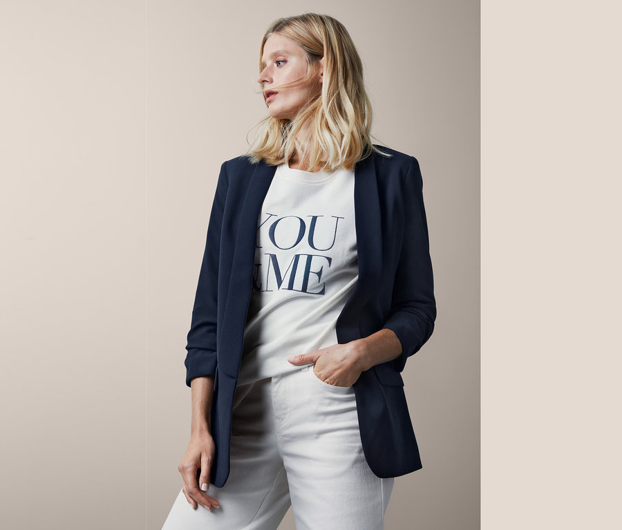 Blonde Frau posiert in einem Blazer mit gerafften Ärmeln und einem weißen T-Shirt mit Aufdruck.