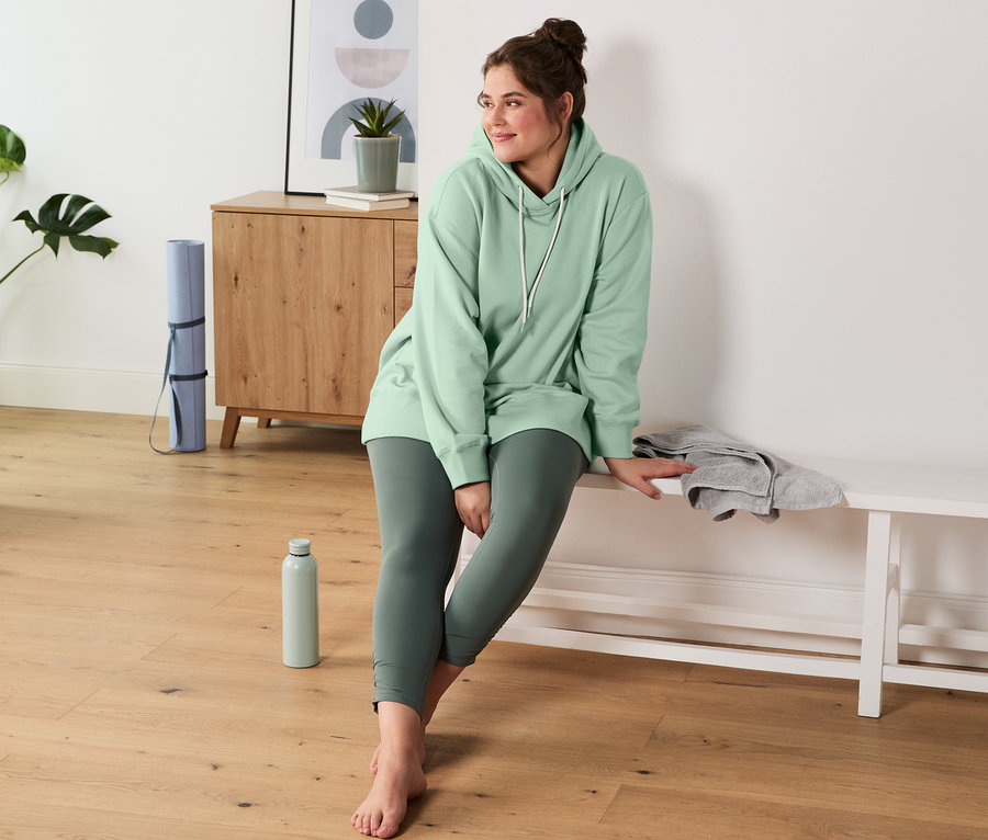 Eine Frau sitzt auf einer Bank und trägt einen mintfarbenen Sweatpullover mit Kapuze und eine Bodyforming-Sporttight.