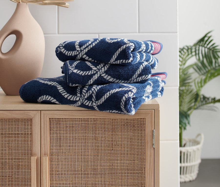 Auf einem Holzschrank liegen gestapelte, marineblaue 2 Handtücher im Maritim-Design mit weißem Seilmuster.