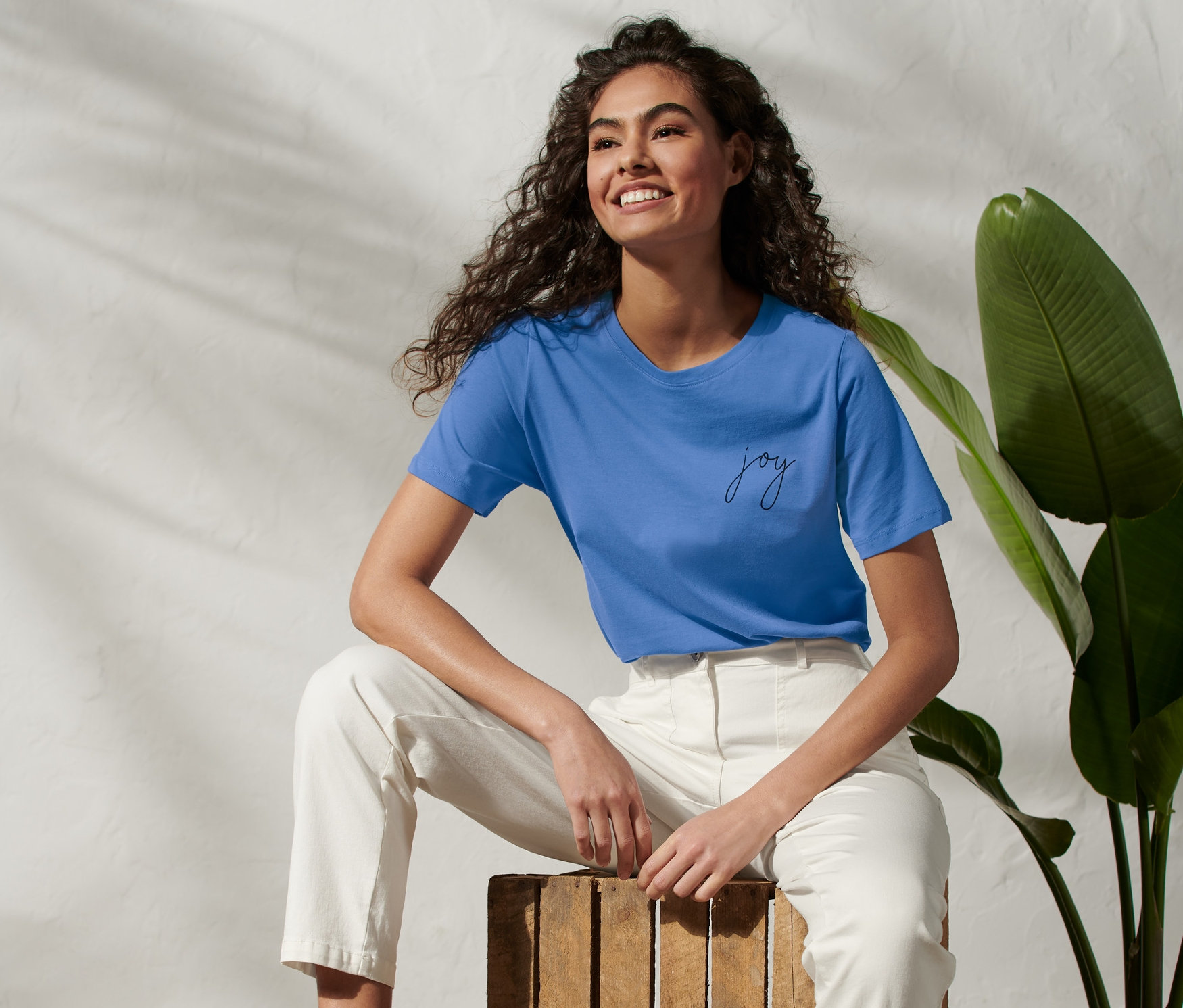 Frau sitzt auf Holzkiste und trägt blaues T-Shirt mit Wording-Print.