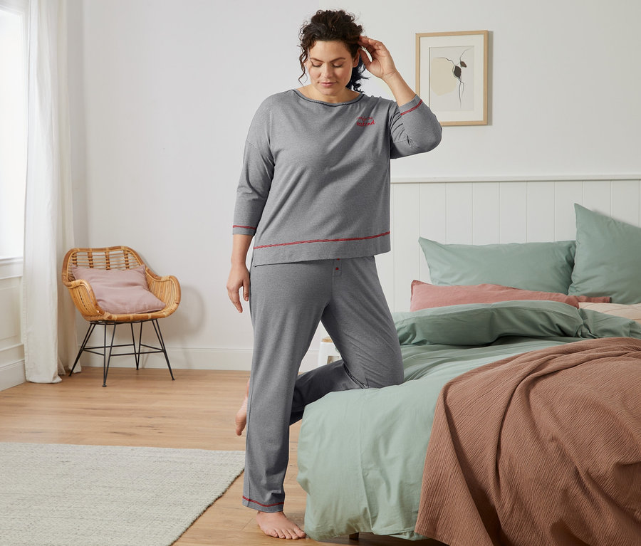 Frau im grauen Pyjama mit feinen Streifen steht am Bett.