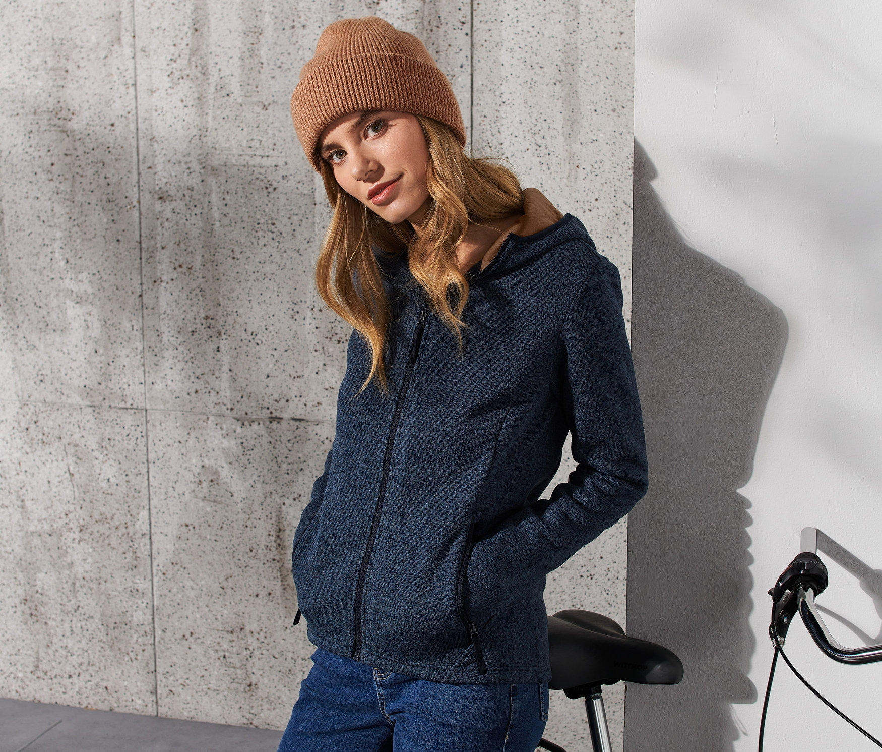 Frau trägt eine dunkelblaue Strickfleece-Jacke und eine braune Mütze, steht neben einem Fahrrad.