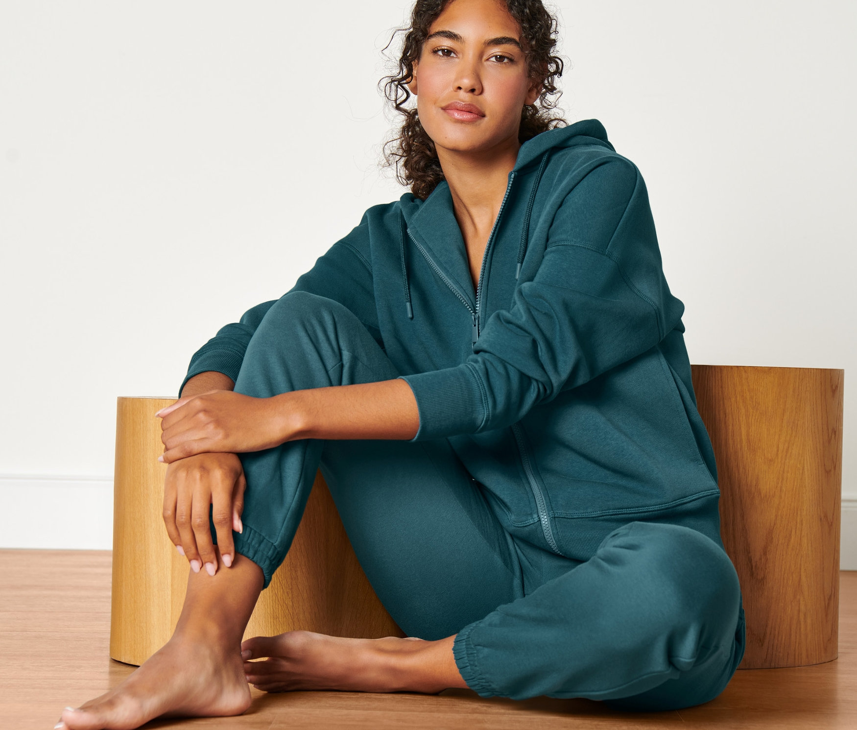 Frau sitzt auf einem Holzhocker und trägt eine dunkelsmaragdgrüne Sweatjacke.
