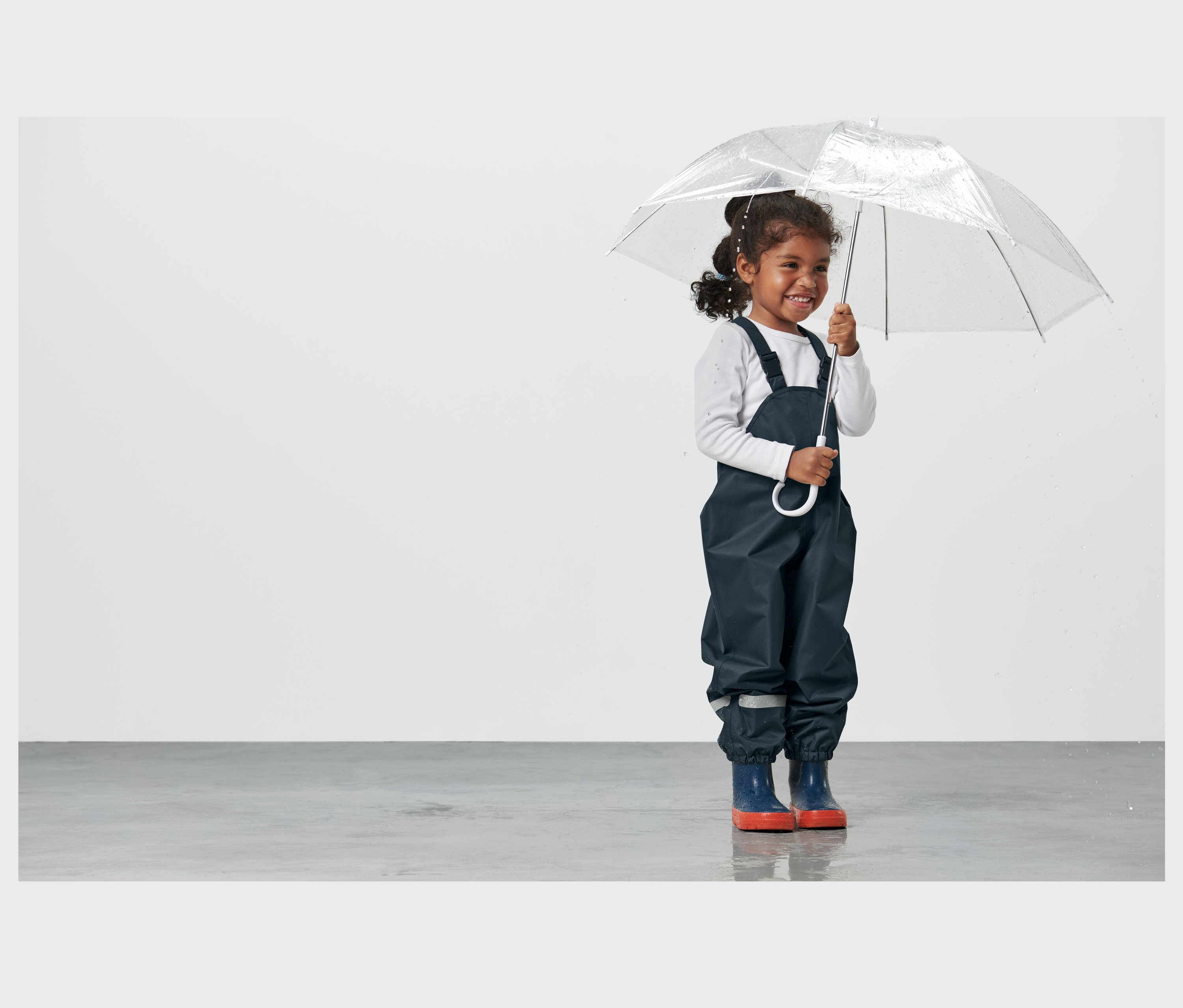 Lächelndes Mädchen hält einen durchsichtigen Regenschirm und trägt eine dunkelblaue Kleinkind-Regenhose.