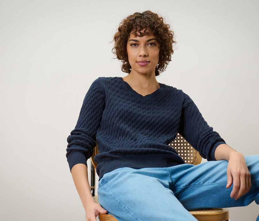 Frau sitzt auf einem Stuhl und trägt einen dunkelblauen Zopfstrickpullover.