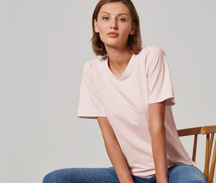 Frau sitzt auf einem Stuhl und trägt ein roséfarbenes Shirt mit Raffung.