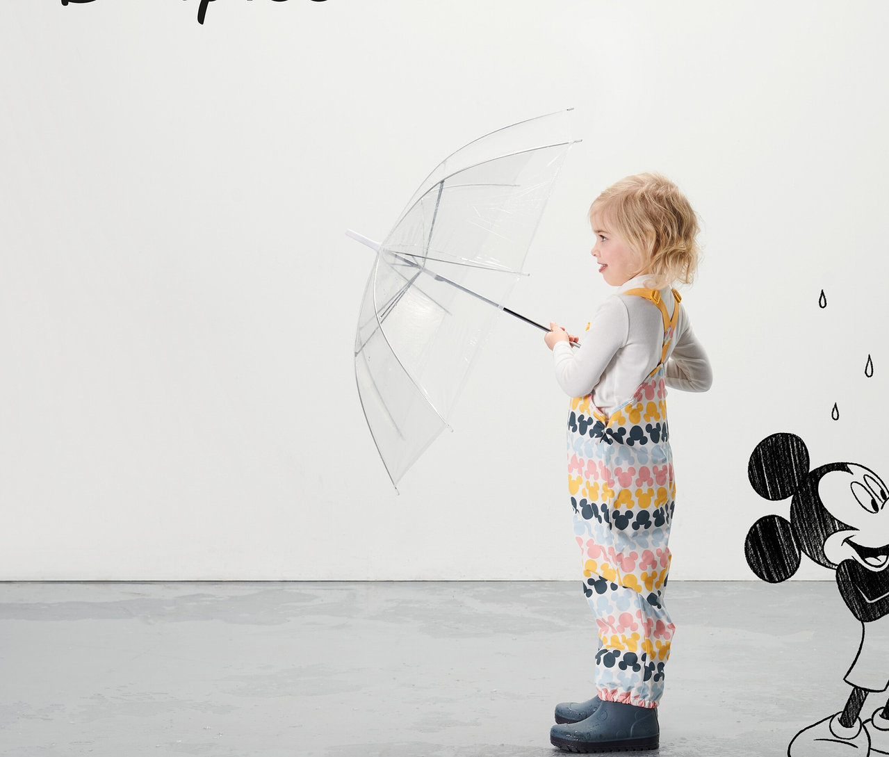 Ein Mädchen hält einen durchsichtigen Regenschirm und trägt eine Kleinkind-Thermo-Regenhose »Micky«, mehrfarbig.