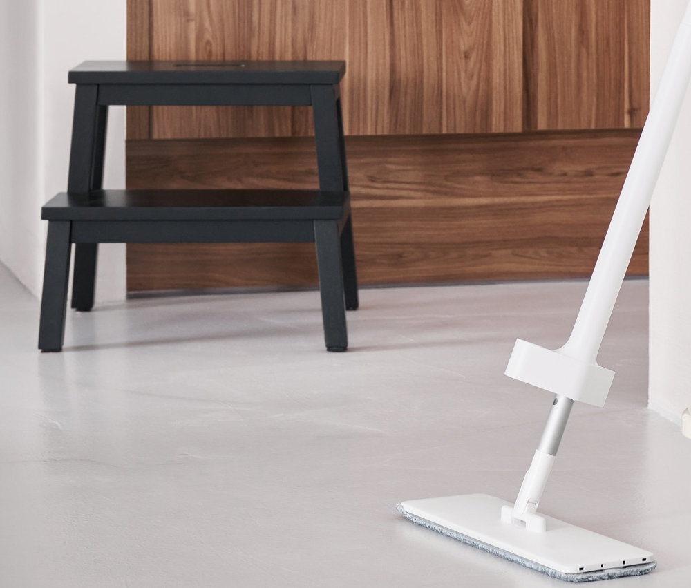 Ein Bodenwischer mit Sprühfunktion steht auf dem Boden, daneben ein faltbarer Putzeimer. Im Hintergrund steht ein Tritthocker mit 2 Stufen.