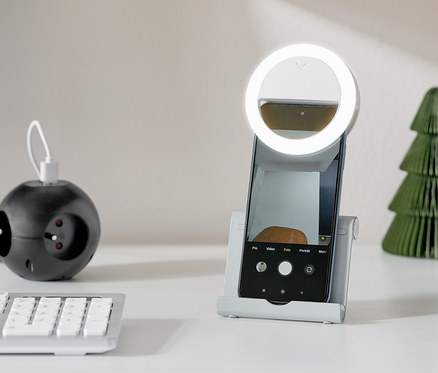 Ein silberner Smartphone-und-Tablet-Ständer hält ein Smartphone mit einem Ringlicht auf einem weißen Tisch.