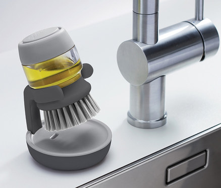 Eine graue Joseph Joseph Spülbürste mit Spülmittelspender steht neben einem Edelstahlspülbecken.