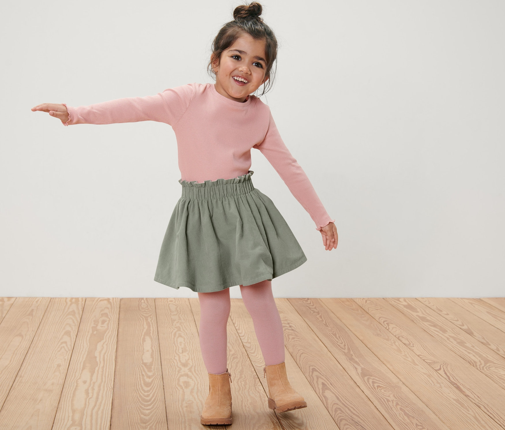 Mädchen mit Dutt tanzt in rosa Shirt, Strumpfhose und khaki Kinder-Cord-Wende-Rock.