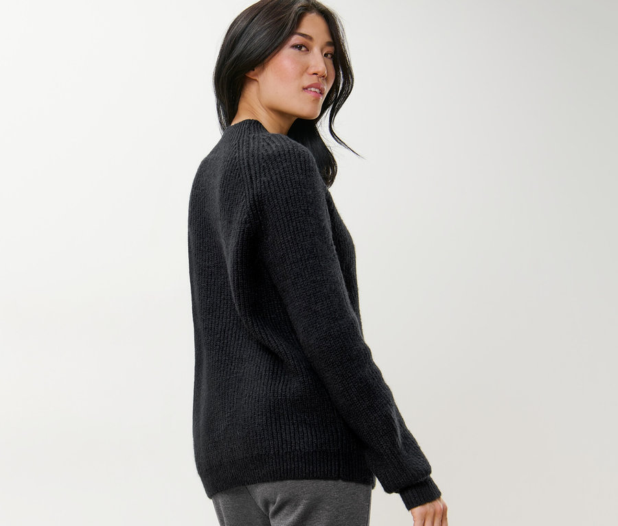Frau blickt über die Schulter und trägt einen schwarzen Grobstrickpullover mit Stehkragen.