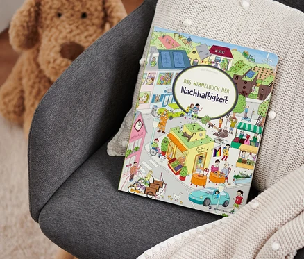 Auf einem grauen Sessel liegen ein beiges Strickplaid und das Buch "Das Wimmelbuch der Nachhaltigkeit". Im Hintergrund ist ein Teddybär zu sehen.