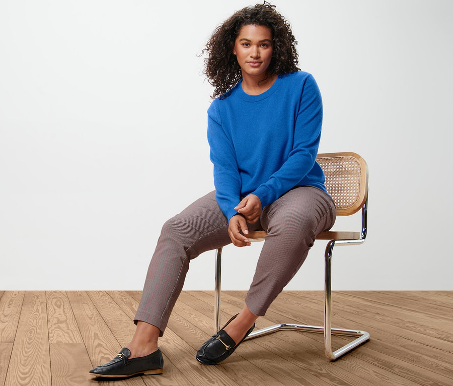Eine Frau sitzt auf einem Stuhl und trägt einen kobaltblauen Feinstrickpullover mit Cashmere und eine karierte Stretchhose.