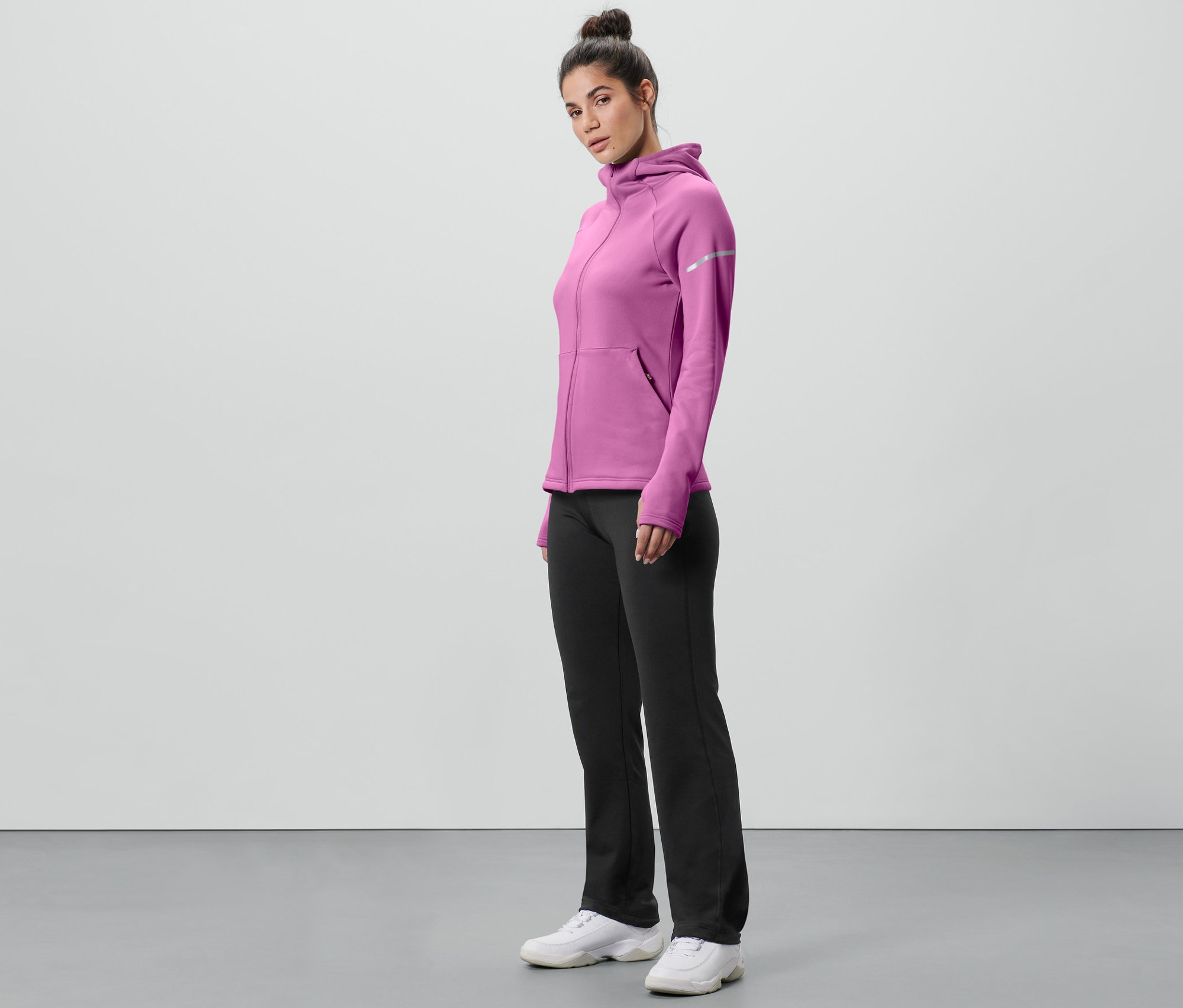 Frau steht seitlich in pinkem Kapuzenpullover und schwarzer Thermohose.