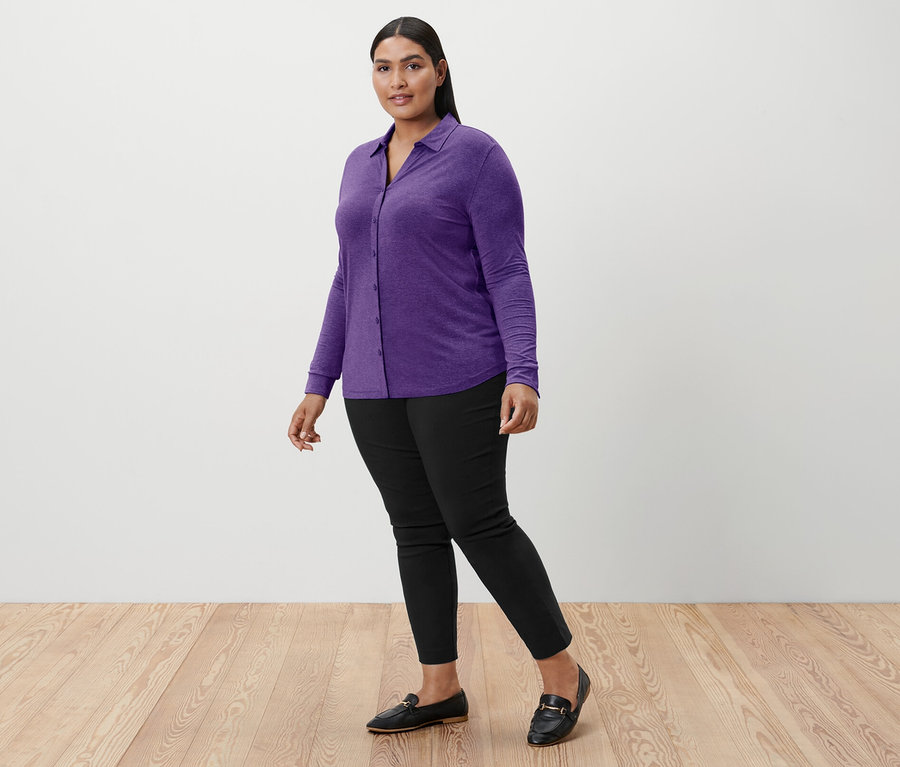 Frau geht, trägt ein lila Blusenshirt mit Knopfleiste, schwarze Hose und schwarze Slipper.