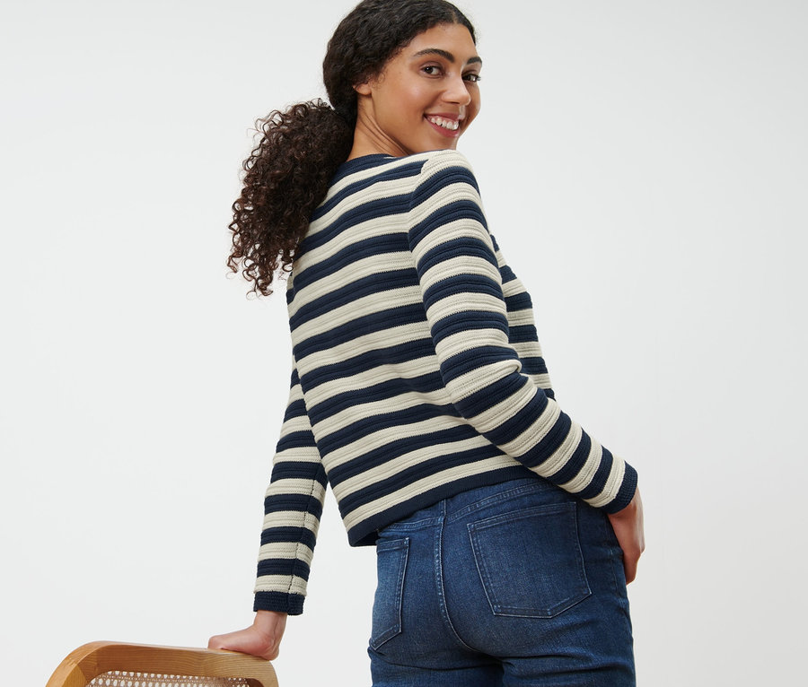 Eine lächelnde Frau blickt über die Schulter und trägt eine Strickjacke aus Strukturstrick und Jeans.