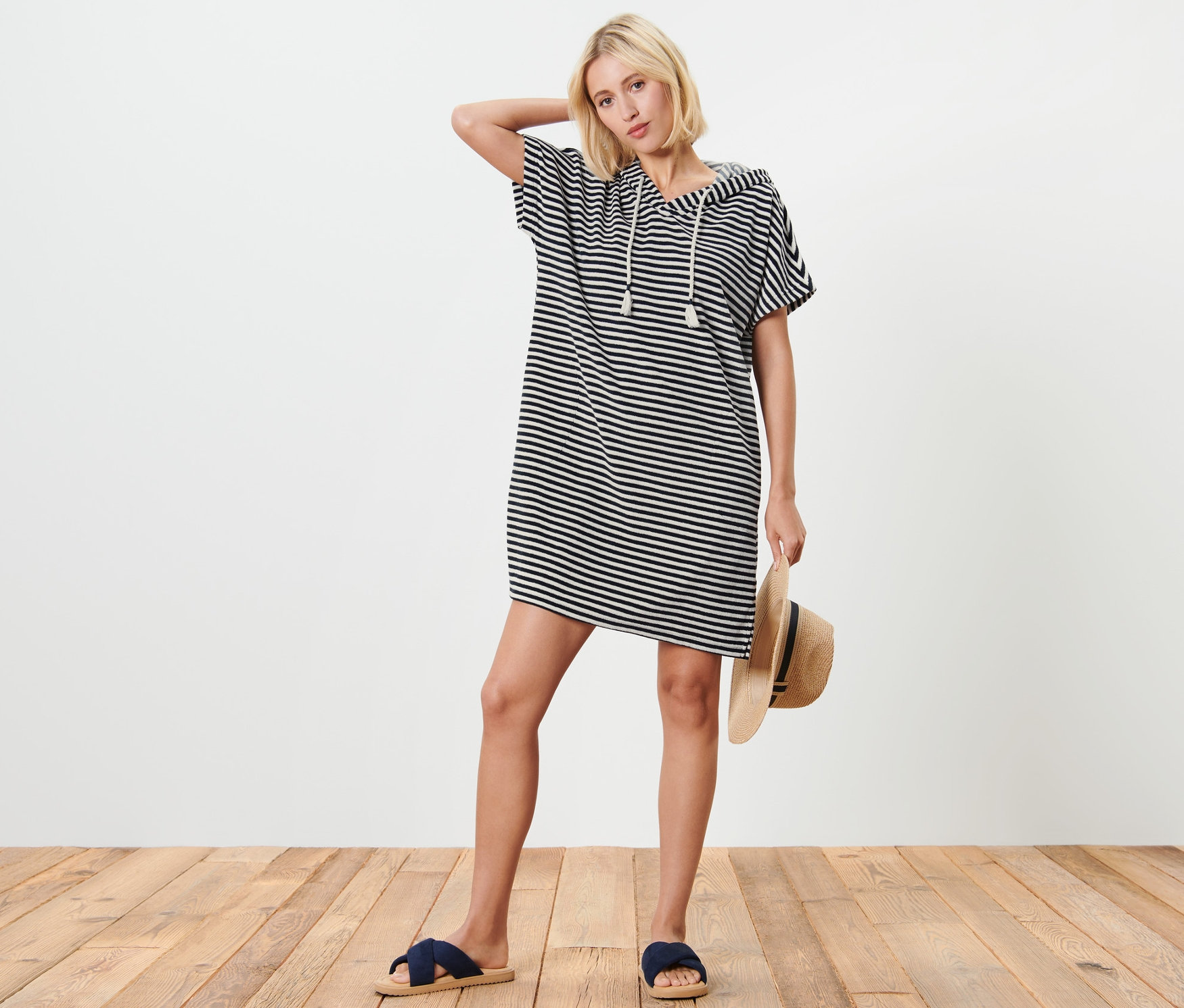 Blonde Frau posiert in blau-weiß gestreiftem Frottee-Strandkleid mit Kapuze, hält einen Strohhut und trägt blaue Pantoletten.