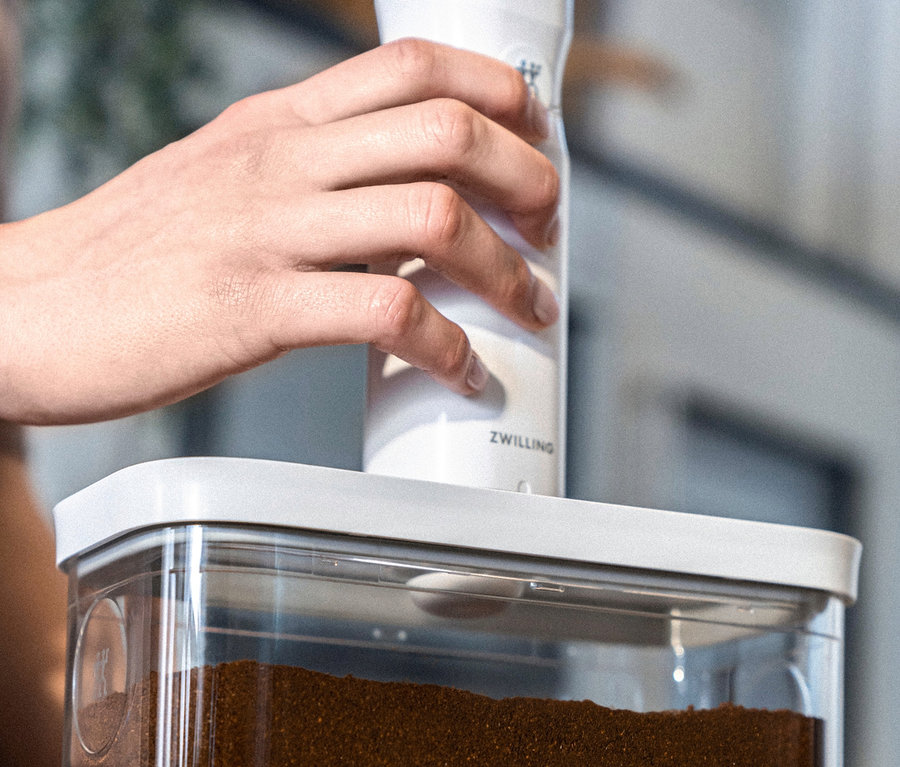 Eine Hand benutzt eine weiße ZWILLING FRESH & SAVE CUBE Vakuumpumpe auf einem durchsichtigen Behälter voller Kaffeepulver.