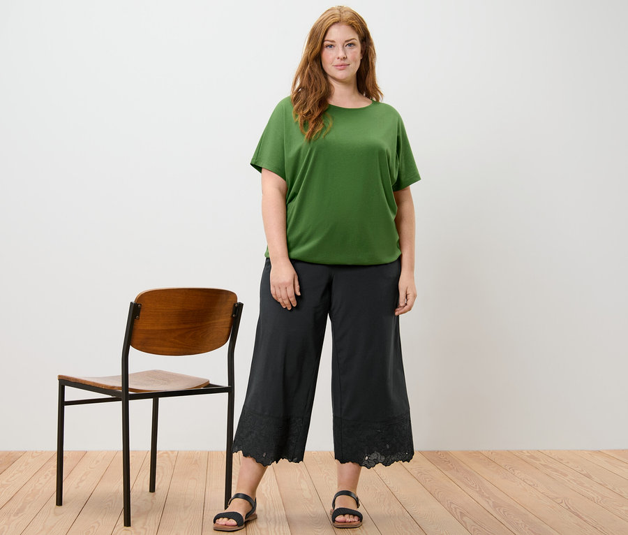 Frau posiert in einem grünen Blusenshirt, einer schwarzen Palazzohose und schwarzen Sandalen. Neben ihr steht ein Holzstuhl.