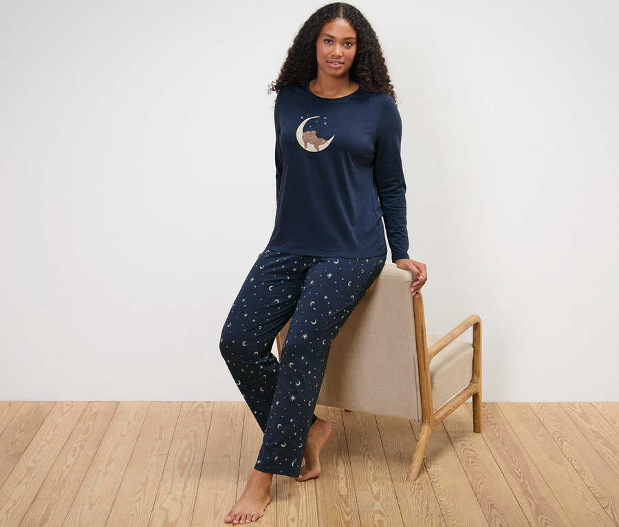 Frau lehnt an Stuhl, trägt dunkelblauen Pyjama mit Mond- und Sternenmuster.