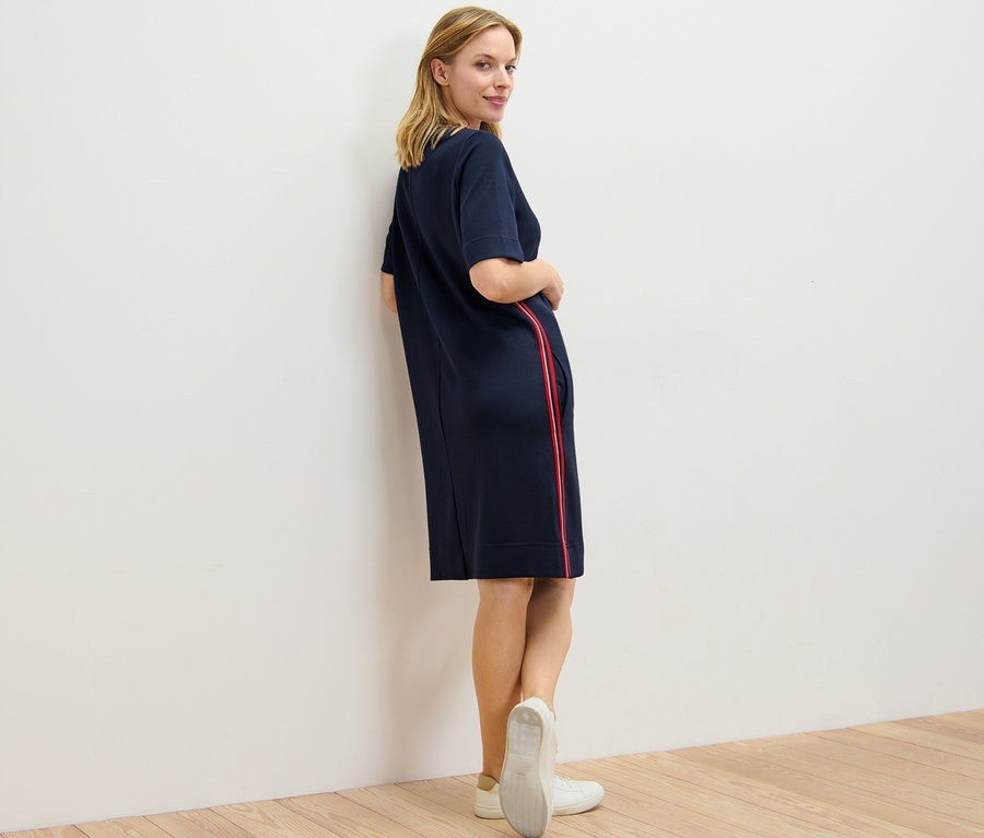 Frau blickt über die Schulter und posiert in einem Sweatkleid mit Galonstreifen.