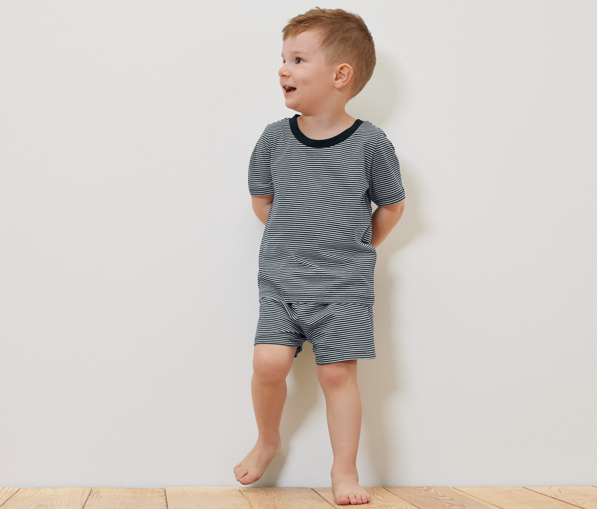 Ein Junge in einem kurzärmeligen 2 Kinder-Pyjama, kurz, steht barfuß auf einem Holzboden neben einem Teddybär.