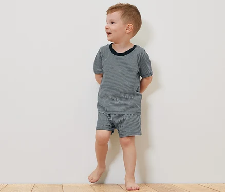 Ein Junge in einem kurzärmeligen 2 Kinder-Pyjama, kurz, steht barfuß auf einem Holzboden neben einem Teddybär.