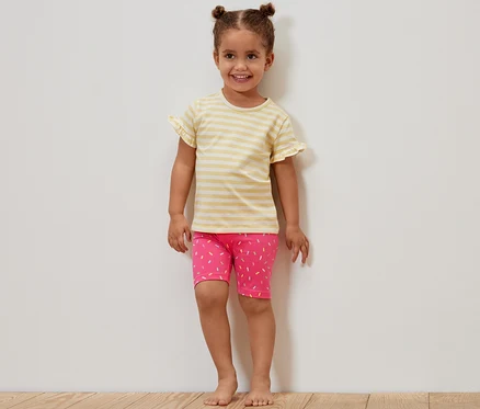 Ein lächelndes Mädchen steht an einer Wand und trägt ein gelb gestreiftes T-Shirt und eine rosa Radlerhose mit Muster. Das Mädchen trägt 2 Kinder-Radler-Leggings, pink und gelb.