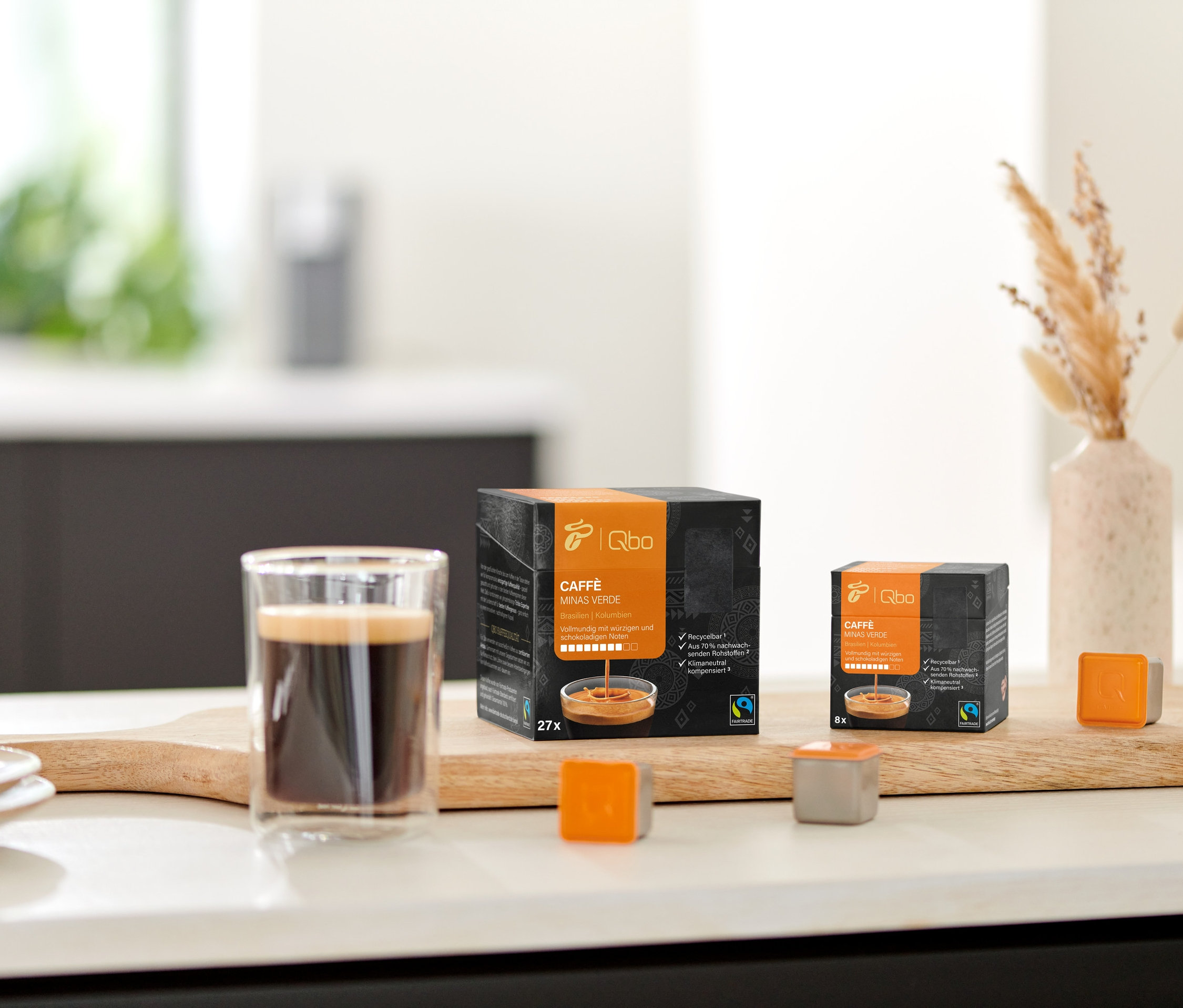 Auf einem Holzbrett stehen ein Glas Kaffee, eine Packung Qbo Caffè MINAS VERDE und einige Kapseln.