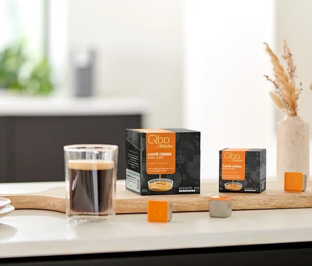 Zwei Qbo Caffè MINAS VERDE Kapselpackungen, ein Glas Kaffee und lose Kapseln auf einem Holzbrett auf der Küchenzeile, daneben Kekse und Trockenblumen.