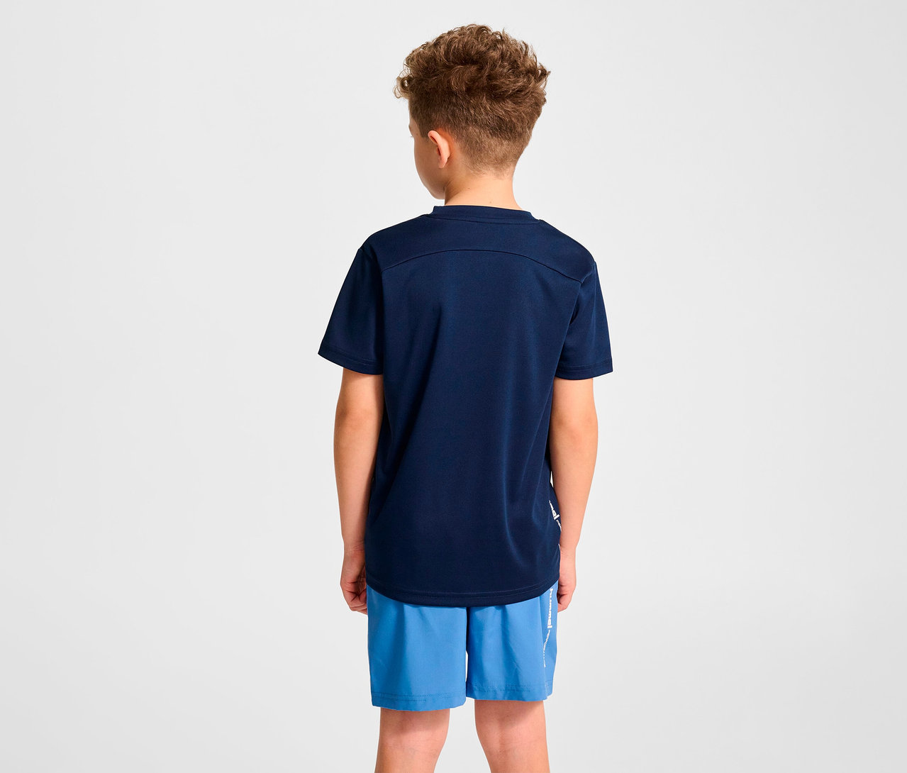 Junge mit dem Rücken zur Kamera trägt ein dunkelblaues HUMMEL HMLJR Pulse Trainings-T-Shirt und blaue Shorts.