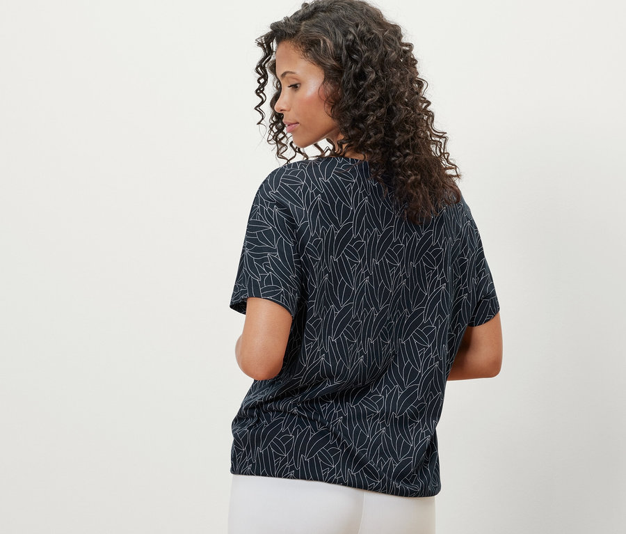 Rückansicht einer Frau mit lockigem Haar, die ein dunkelblaues, überschnittenes Blusenshirt und weiße Hosen trägt.