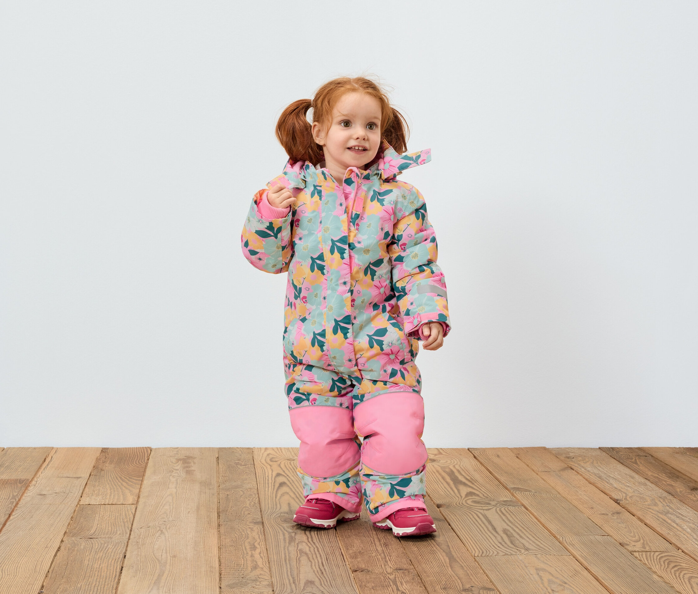 Ein rothaariges Mädchen im Blumenoverall und rosa Schuhen steht auf einem Holzboden vor einer weißen Wand.