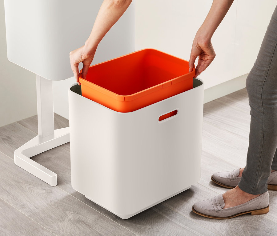 Eine Person setzt einen orangefarbenen Behälter in einen weißen Abfalleimer.