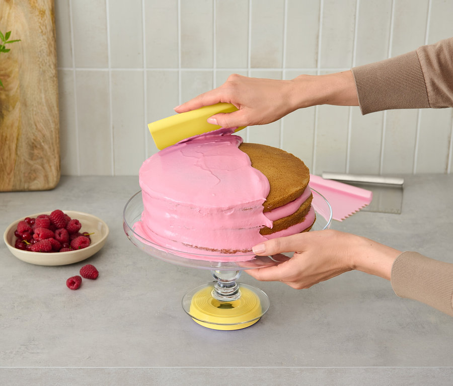 Eine Person verziert einen rosa Kuchen auf einem Glasständer mit einem gelben Werkzeug.