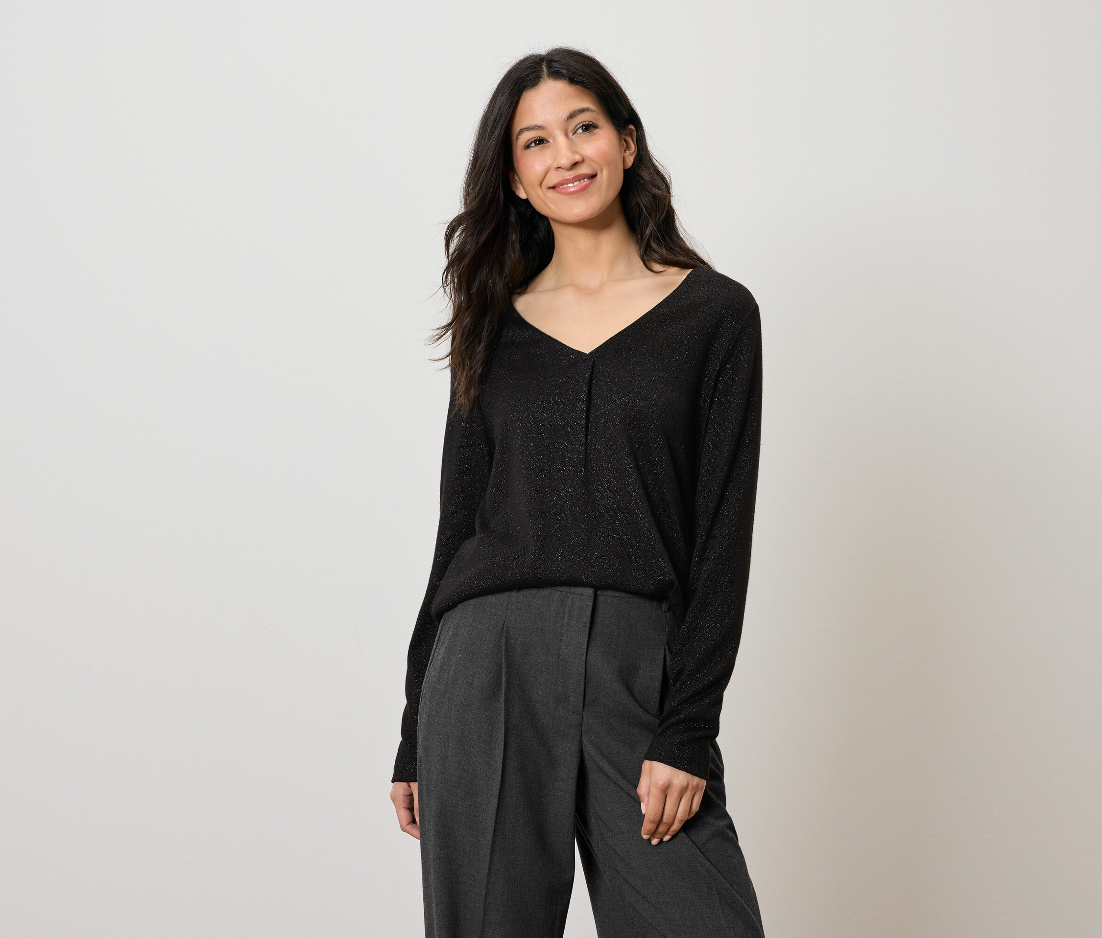 Frau mit langen dunklen Haaren trägt ein schwarzes Langarmshirt und eine graue Hose.