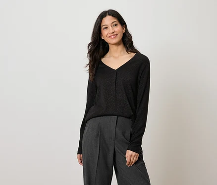 Frau mit langen dunklen Haaren trägt ein schwarzes Langarmshirt und eine graue Hose.