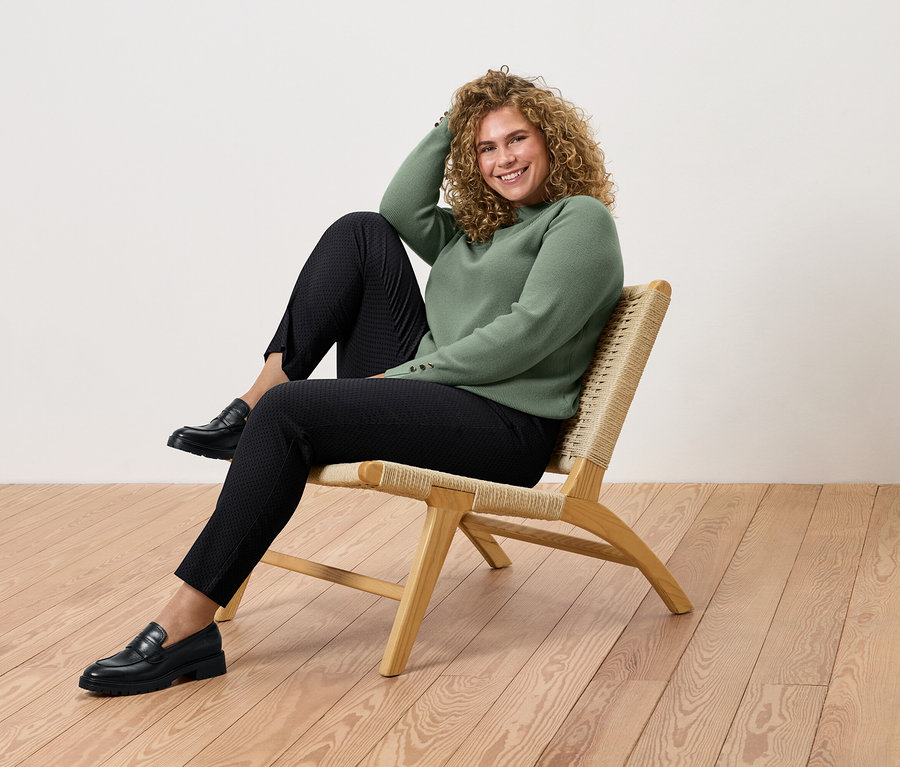 Frau mit lockigem Haar sitzt auf einem Rattansessel, trägt einen grünen Pullover, schwarze Hosen und Schuhe.