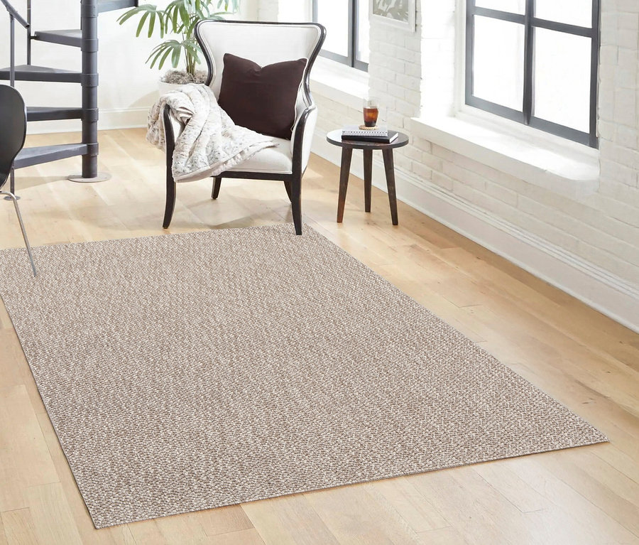 Ein beiger Teppich »Twine« mit weichem Chevron-Muster liegt auf einem Holzboden im Wohnzimmer. Im Hintergrund sind ein Sessel, ein Tisch und ein Treppenhaus zu sehen.