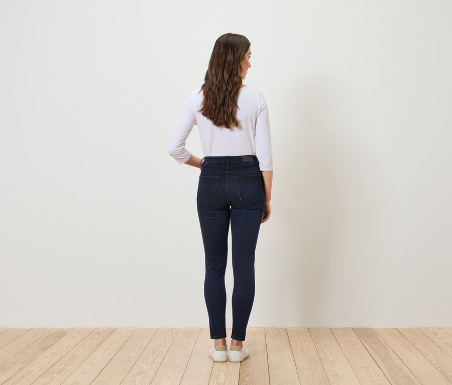 Von hinten ist eine Frau mit braunem, gewelltem Haar in blauen Jeans und einem weißen T-Shirt zu sehen.