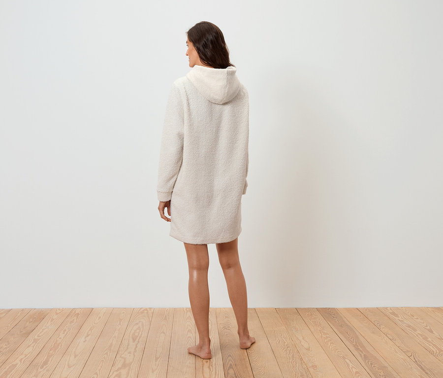 Eine Frau in einem cremefarbenen Kapuzenkleid steht mit dem Rücken zur Kamera auf einem Holzboden.